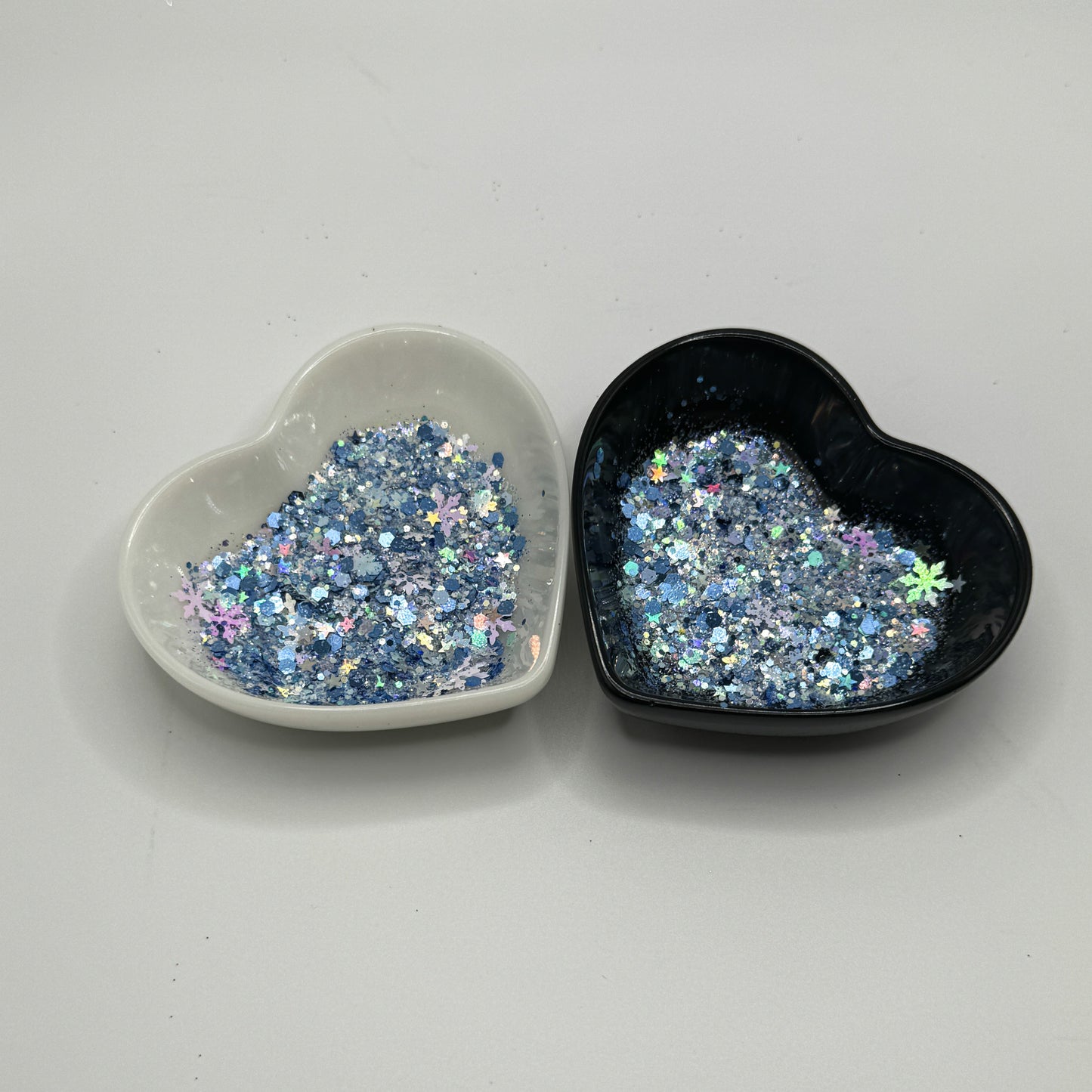 Two heart shaped containers, one black, one white, containing a Blue/White/Rainbow/Silver chunky glitter mix with hexagons, snowflakes and stars.