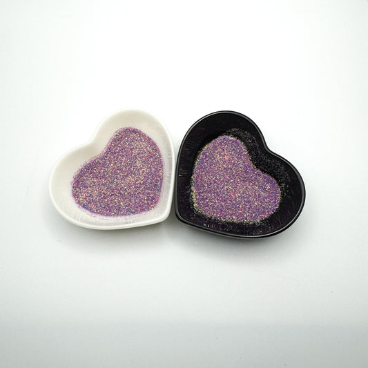 Two heart-shaped dishes containing extra fine glitter, one white and one black, with the glitter appearing purple in color.