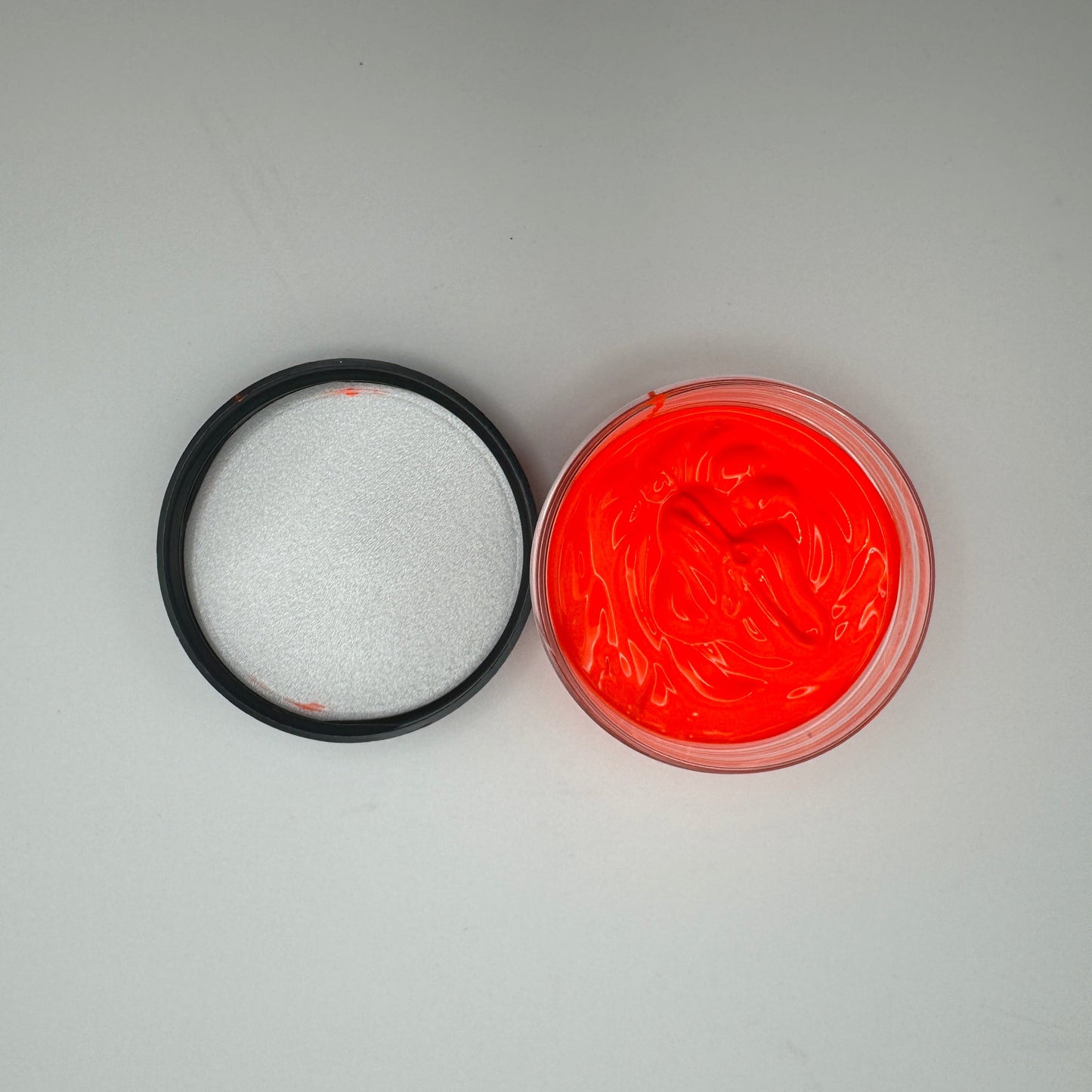 A jar of coral pigment paste is open with its lid beside it on a white surface. The pigment paste is bright coral, and the lid is black.