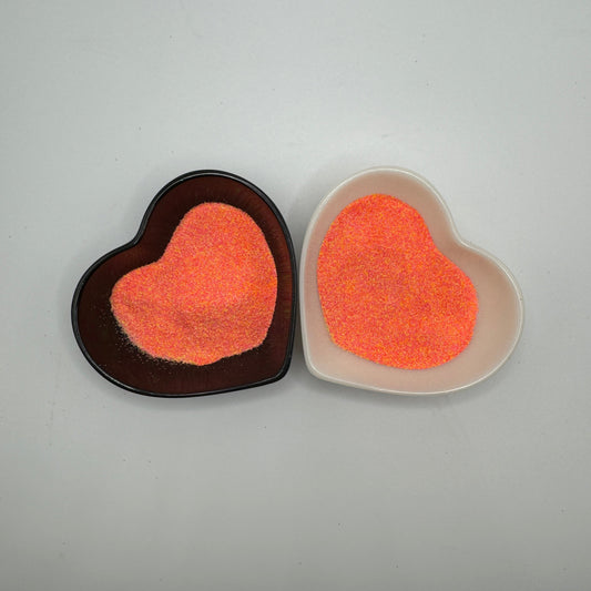 Two heart-shaped samples of orange and pink glitter, one placed on a dark background and the other on a light background, showcasing the color and texture of the glitter.
