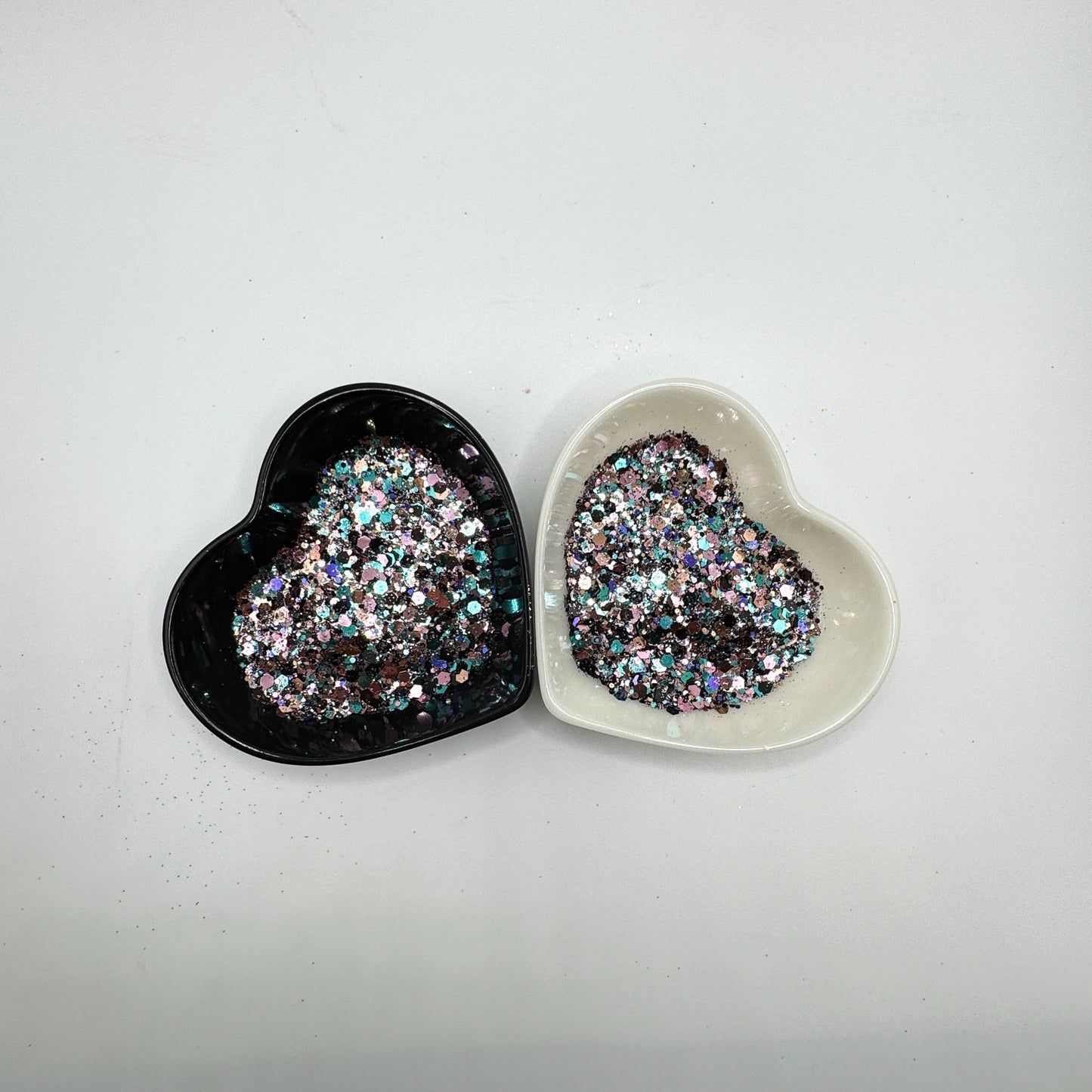 Two heart shaped containers, one black, one white, containing a Blue/Black/Light Teal/Rose Gold/Light Pink chunky glitter mix.