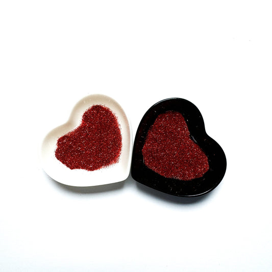 Two heart-shaped containers of glitter, one black and the other white, both showing an extra fine, red glitter mix.