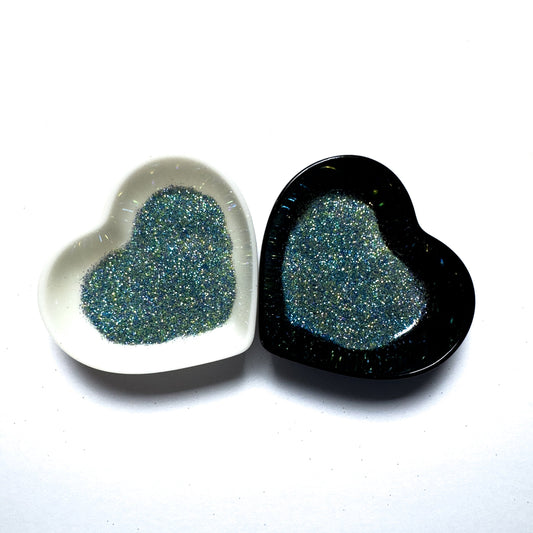 Two heart-shaped containers of glitter, one black and the other white, both showing an extra fine, blue, yellow, green glitter mix.