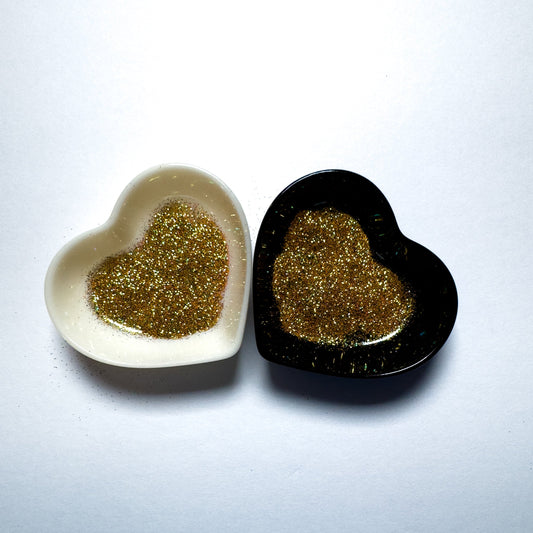 Two heart-shaped containers of glitter, one black and the other white, both showing an extra fine, gold glitter mix.