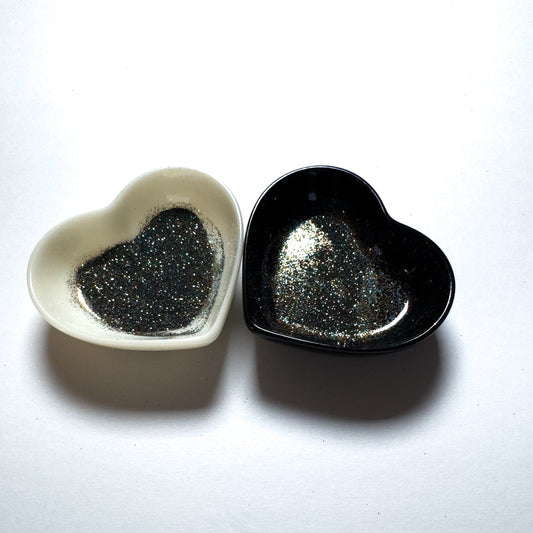 Two heart-shaped containers of glitter, one black and the other white, both showing an extra fine, multicolored glitter mix.