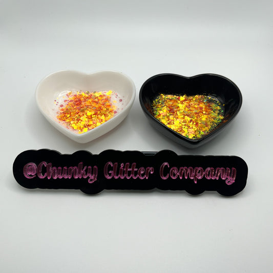 Two heart shaped containers, one black, one white, containing a Orange/Burnt Orange chunky glitter mix, with a label in front that reads '@ChunkyGlitterCompany'.