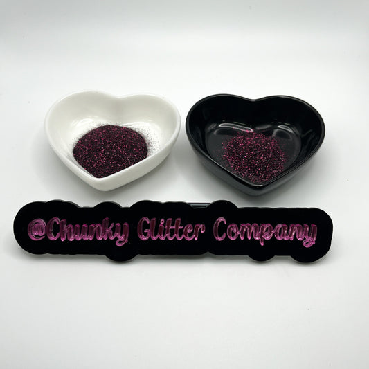 Two heart shaped containers, one black, one white, containing a dark extra fine glitter mix, with a label in front that reads '@ChunkyGlitterCompany'.