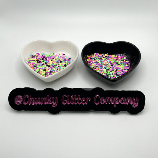 Two heart shaped containers, one black, one white, containing a Pink/Yellow/Blue/Green/Purple chunky glitter mix, with a label in front that reads '@ChunkyGlitterCompany'.