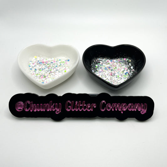 Two heart shaped containers, one black, one white, containing a White/Rainbow chunky glitter mix, with a label in front that reads '@ChunkyGlitterCompany'.