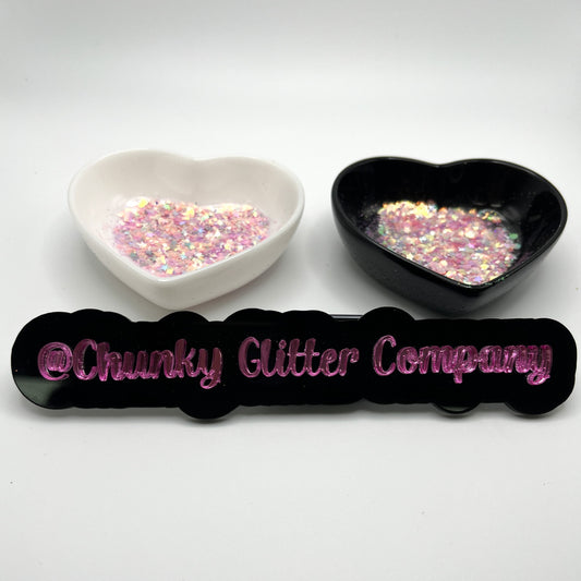 Two heart shaped containers, one black, one white, containing a Purple/Orange/White/Silver chunky glitter mix with hexagons and stars with a label in front that reads '@ChunkyGlitterCompany'.