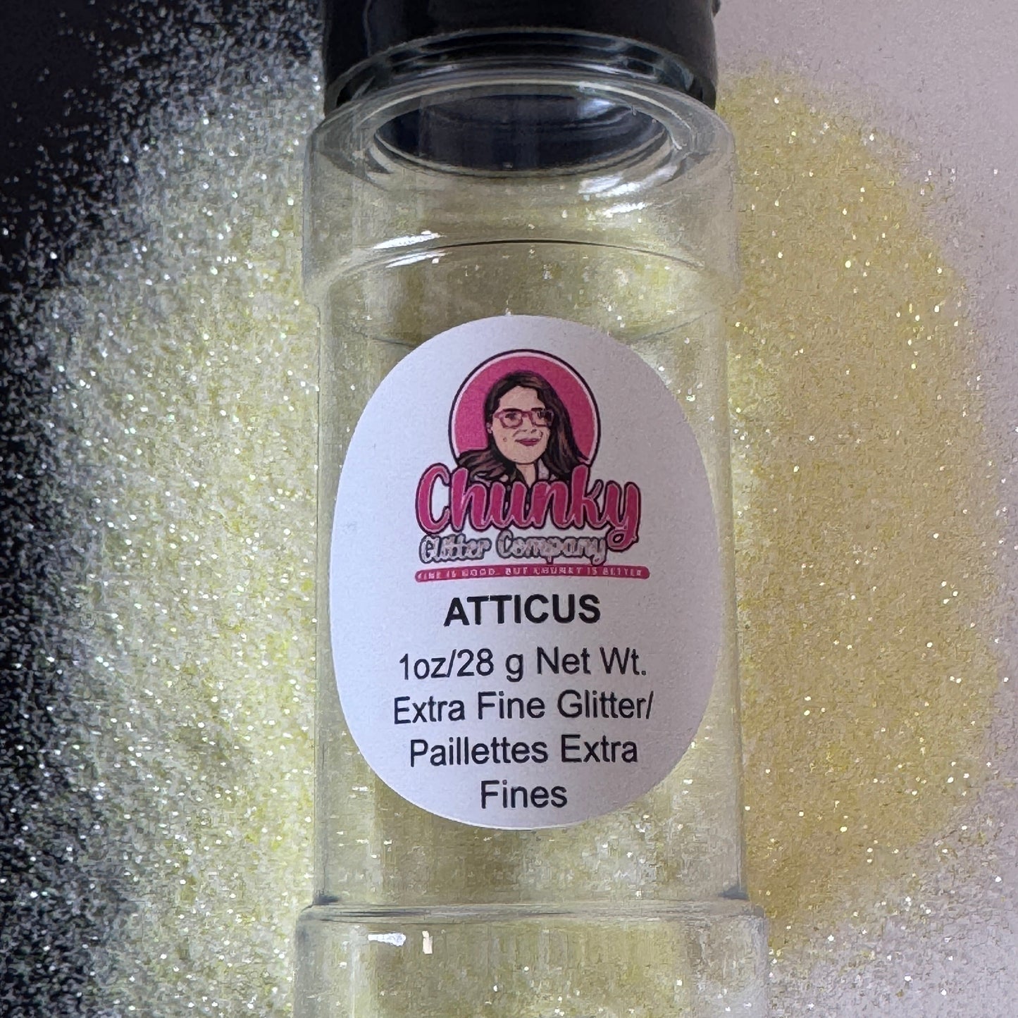 A clear plastic jar containing extra fine pastel yellow glitter with a label on the front showcasing the brand and product name, 'Atticus'.