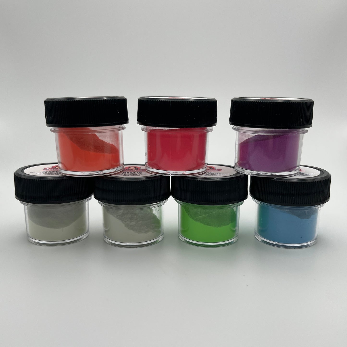 7 small jars of various colours of glow in the dark powders on a white background