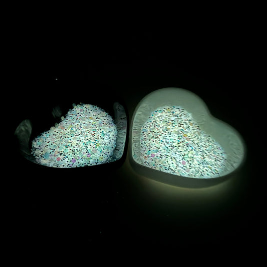 A photograph taken in the dark of two heart shaped containers, one black, one white, containing a White/Rainbow glow in the dark chunky glitter mix.