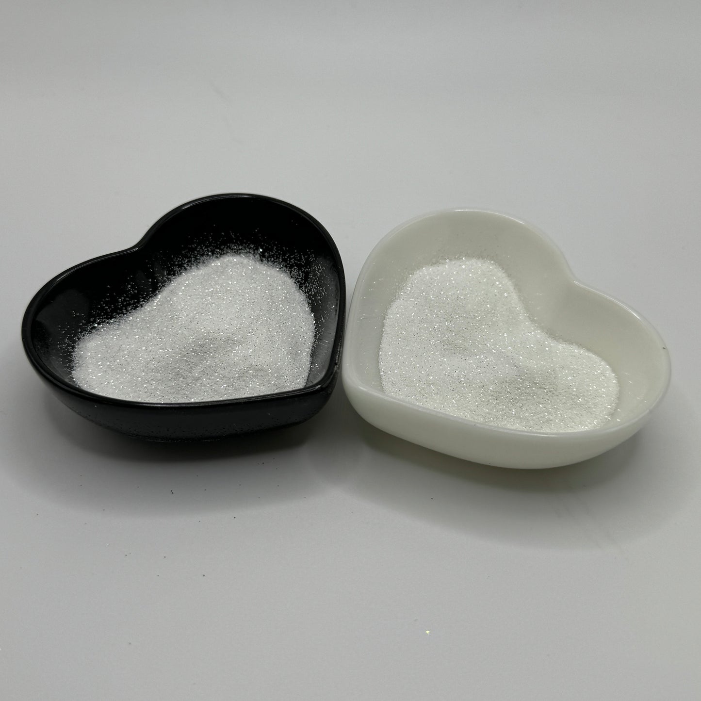 Two heart-shaped containers of glitter, one black and the other white, both showing an extra fine, white glitter.