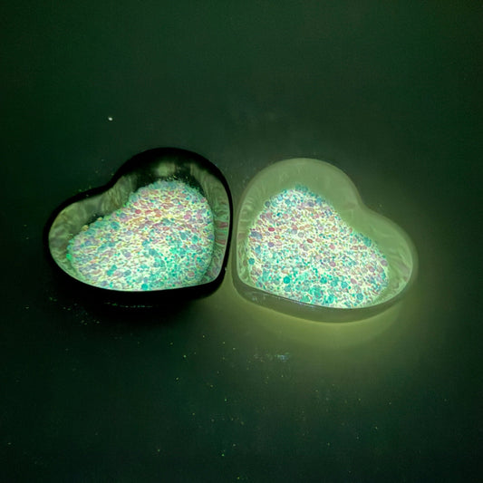 A photograph taken in the dark of two heart shaped containers, one black, one white, containing a White/Rainbow glow in the dark chunky glitter mix.