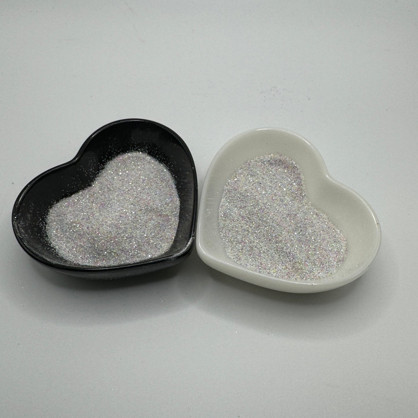 Two heart-shaped containers of glitter, one black and the other white, both showing an extra fine, white rainbow glitter mix