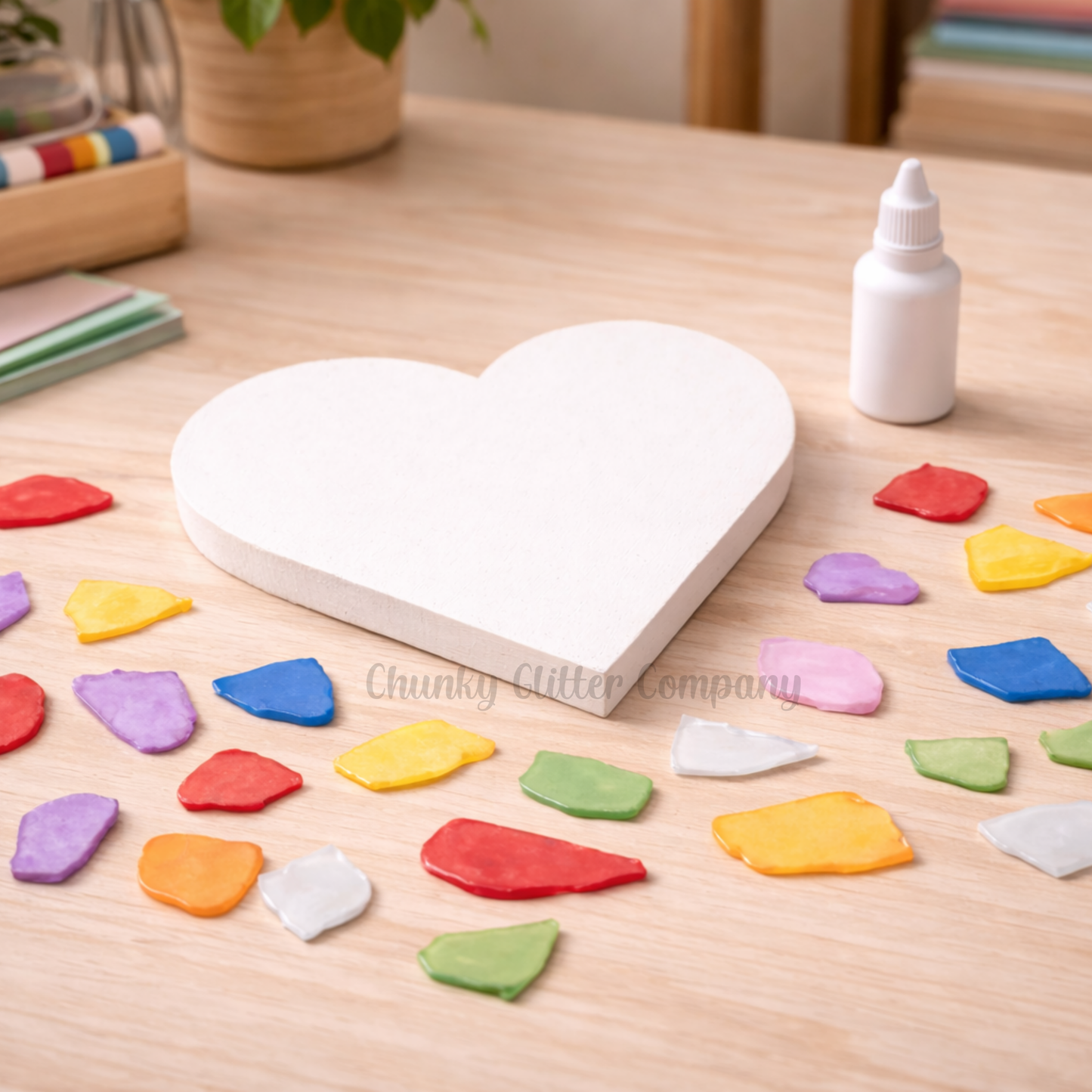 Heart-shaped white board with colorful beach glass on a wooden table, surrounded by a bottle of glue and a plant.