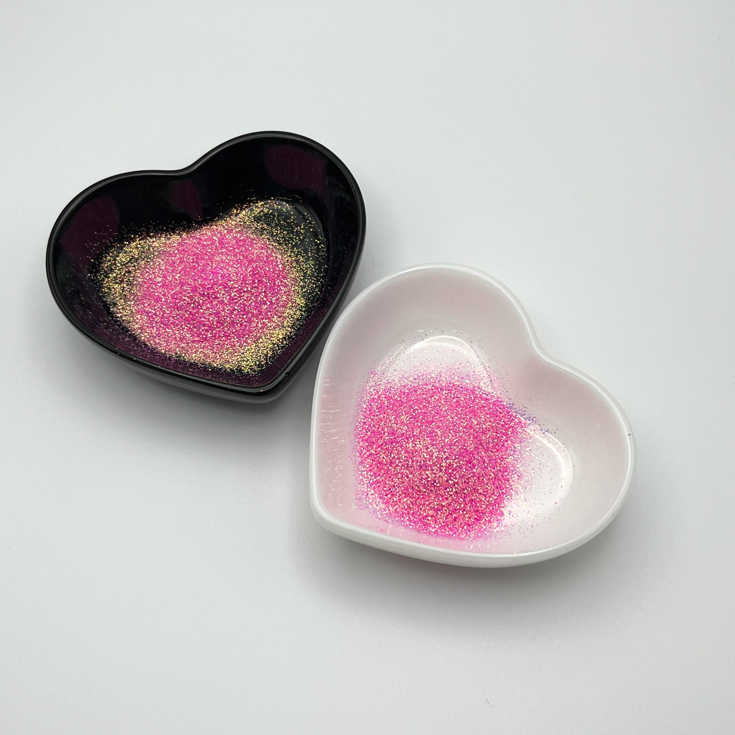 Two heart-shaped dishes, one black and one white, both containing light pink extra fine glitter.