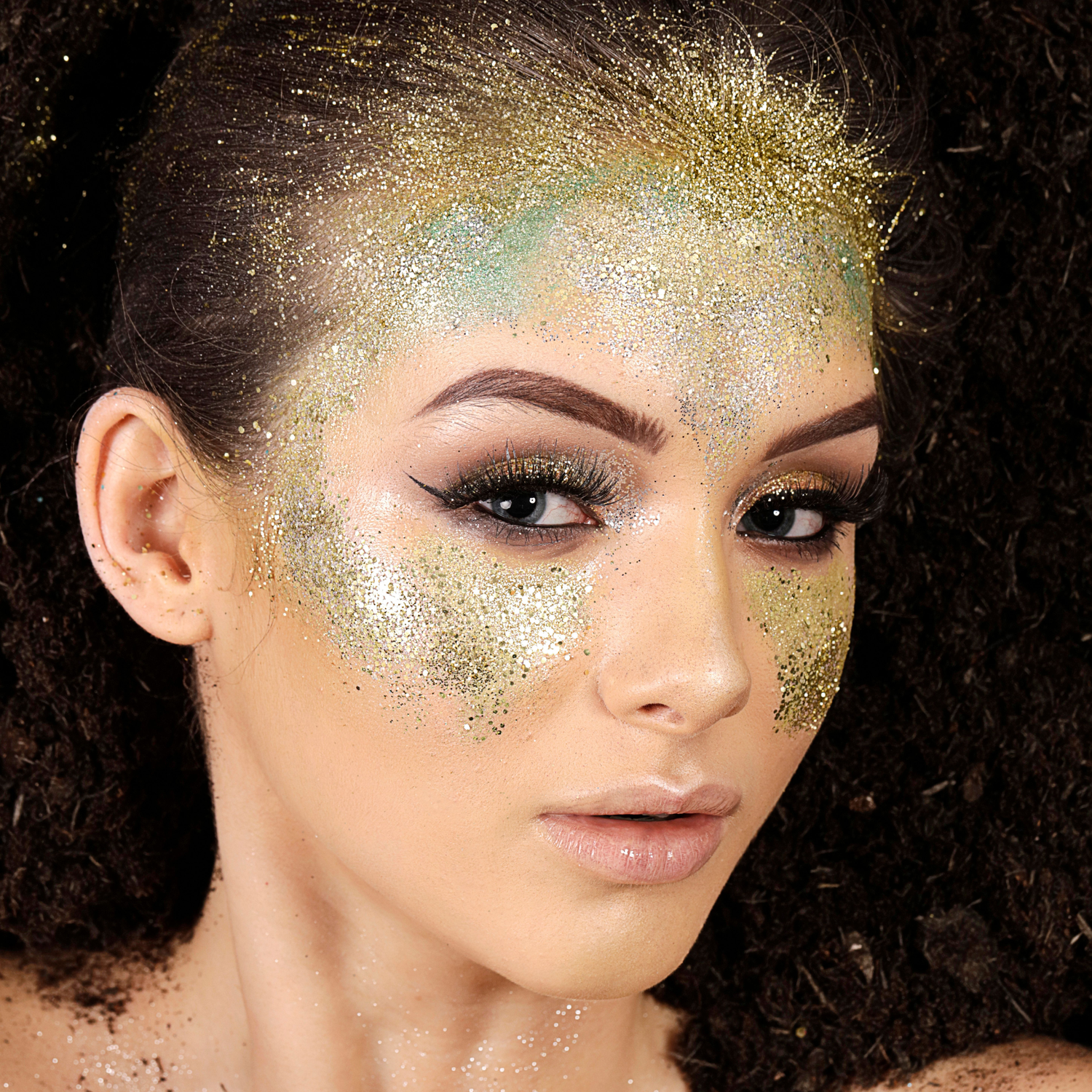 A model face surrounded by dirt with her makeup done and green and gold glitter on her face.