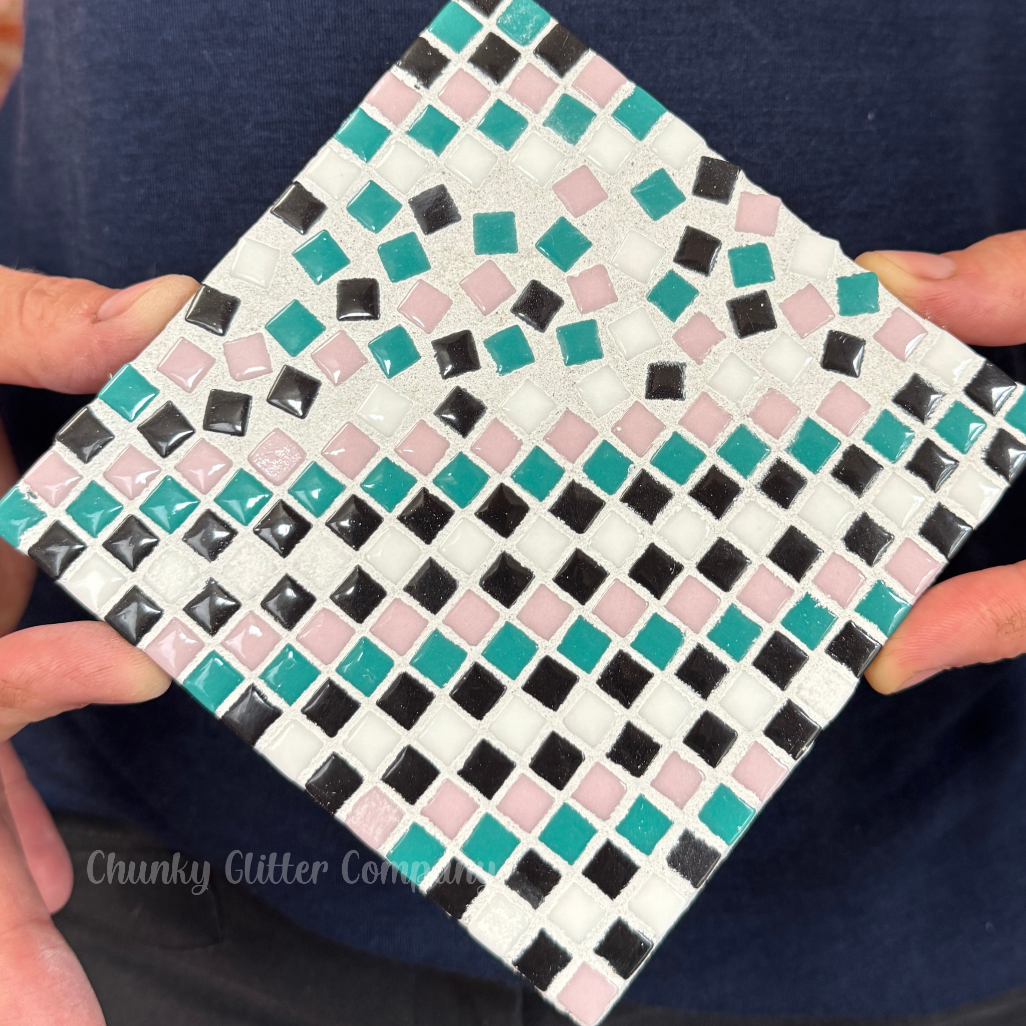 Small square tile with a mosaic pattern held by hands against a dark background