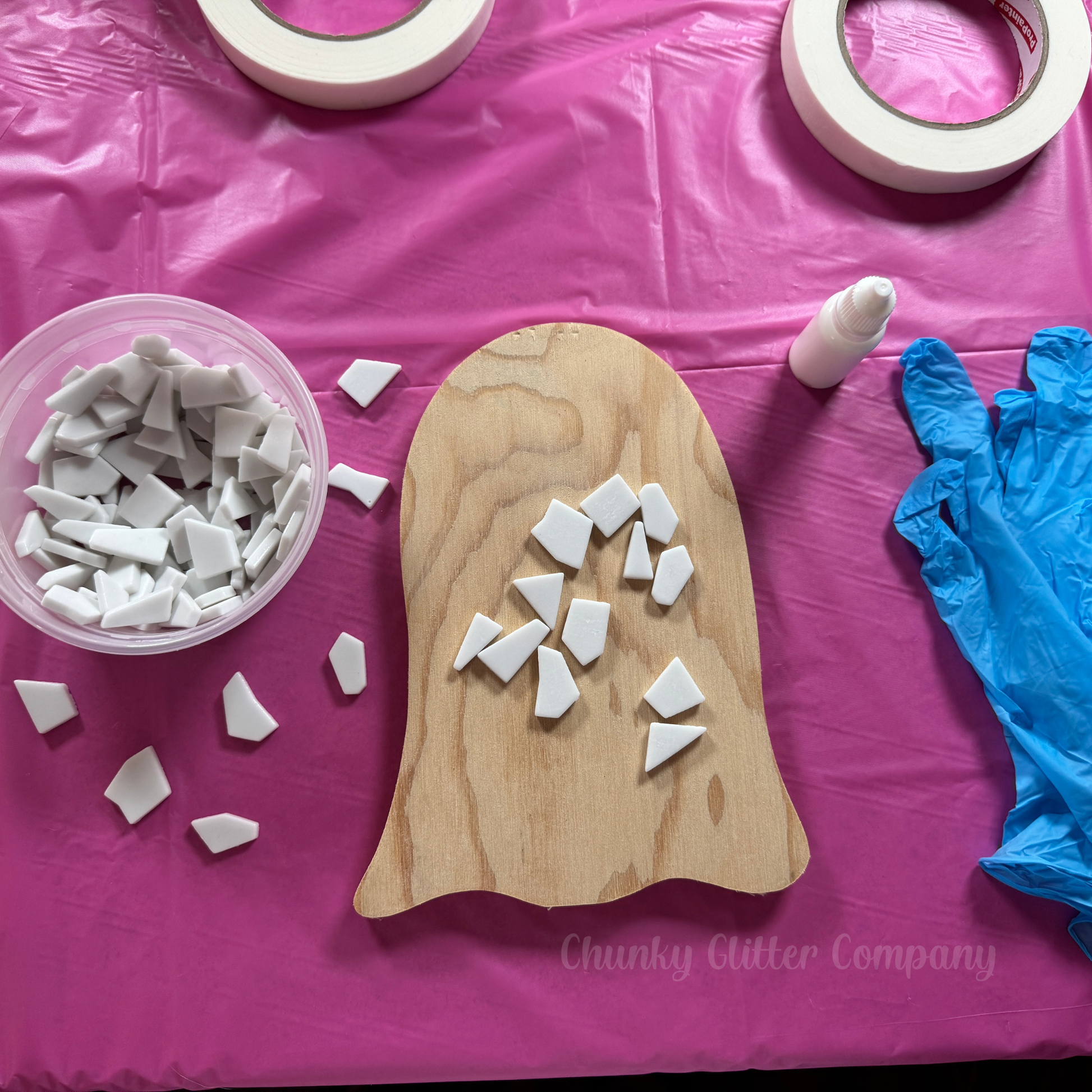 Wooden ghost shaped board with ceramic pieces on a pink table cloth, surrounded by tape, gloves, and a container of more ceramic pieces.