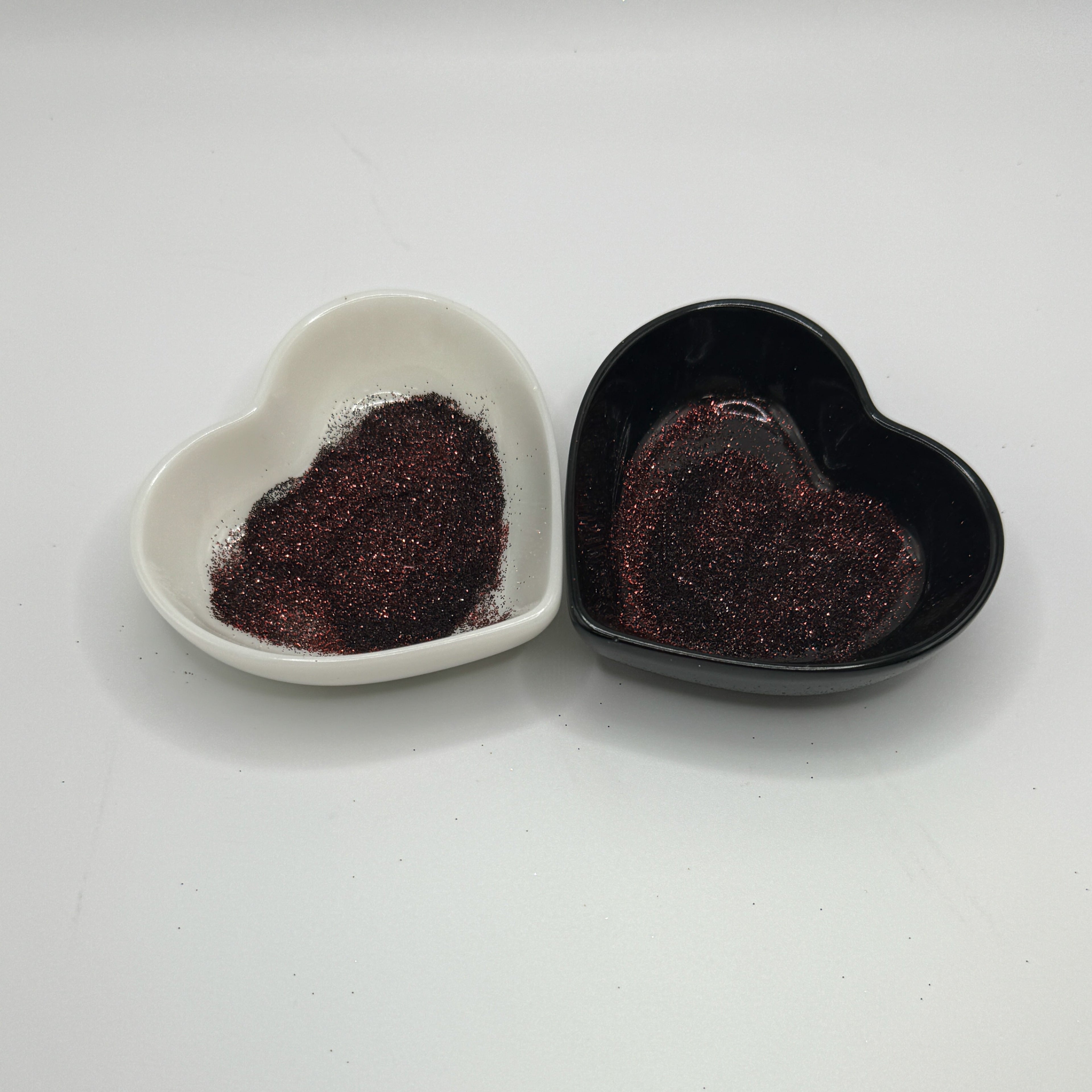 Two heart shaped containers, one black, one white, containing a dark brown extra fine glitter mix.