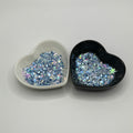 Two heart shaped containers, one black, one white, containing a Blue/White/Rainbow/Silver chunky glitter mix with hexagons, snowflakes and stars.