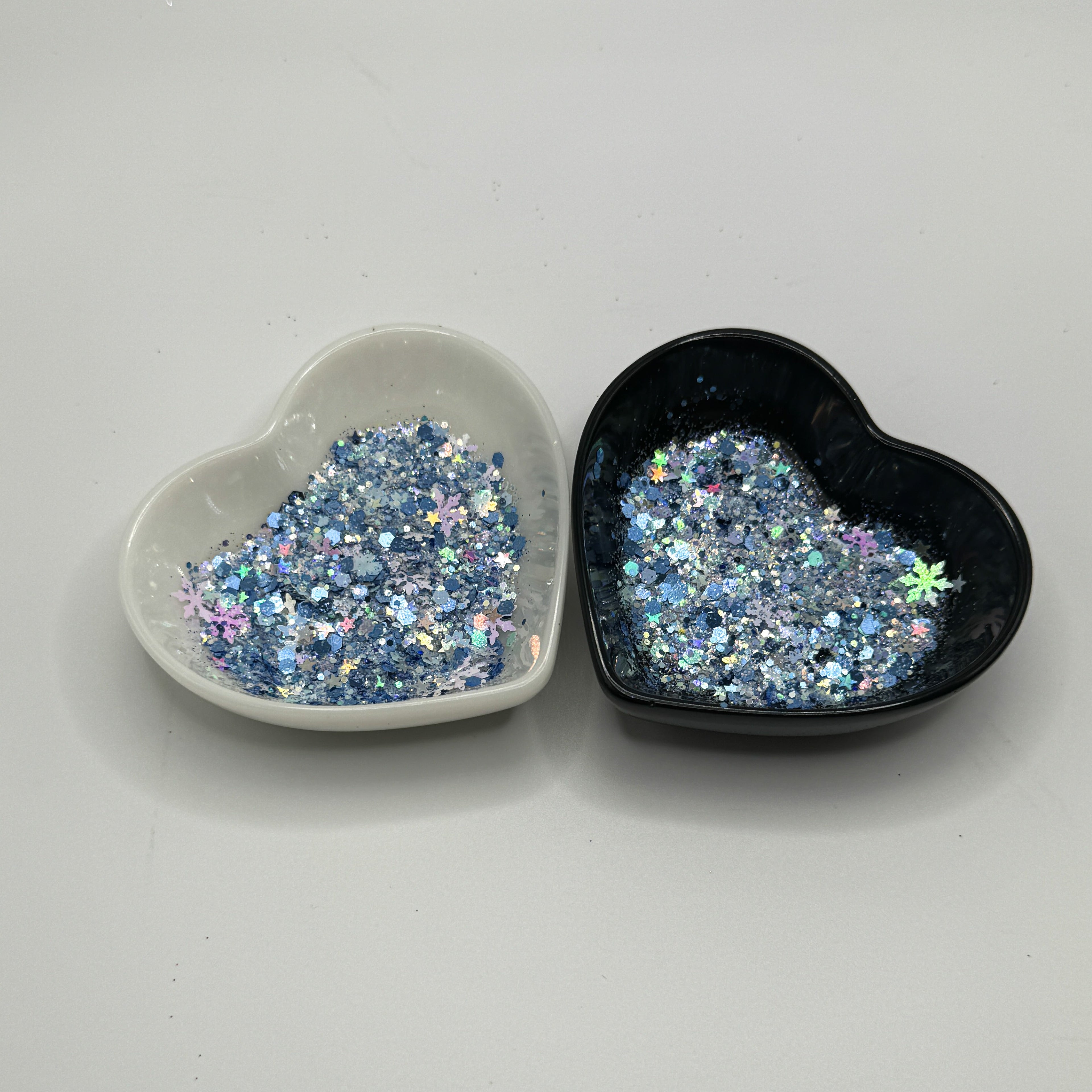 Two heart shaped containers, one black, one white, containing a Blue/White/Rainbow/Silver chunky glitter mix with hexagons, snowflakes and stars.