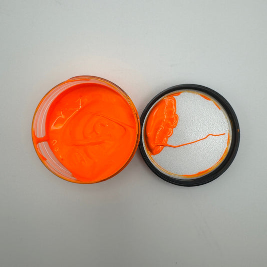 A jar of orange pigment paste is open with its lid beside it on a white background.