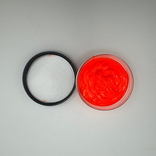 A jar of coral pigment paste is open with its lid beside it on a white surface. The pigment paste is bright coral, and the lid is black.
