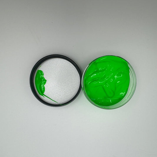A jar of electric green pigment paste, opened and showing the green-coloured product inside. The lid is placed next to it on the white surface.