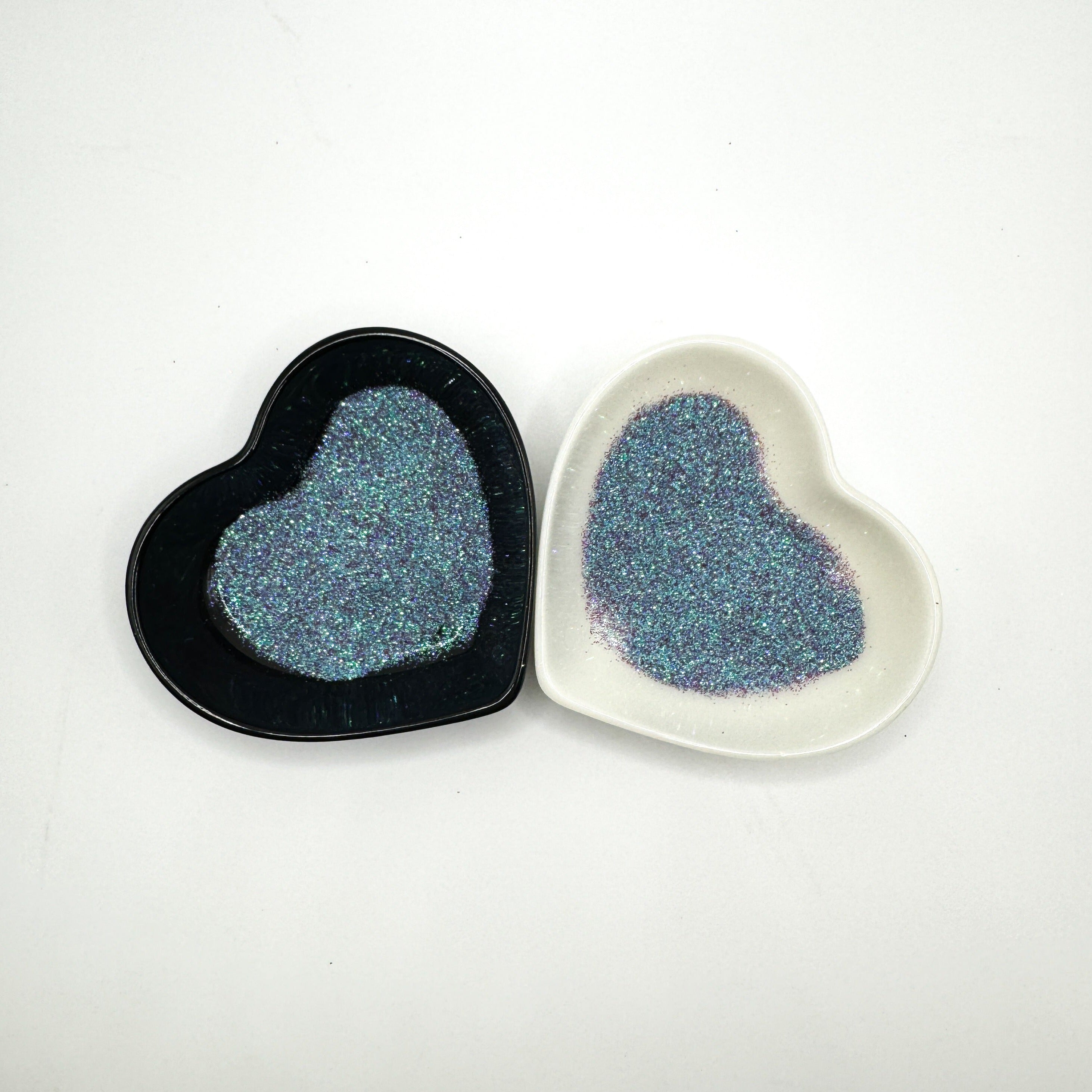 Two heart-shaped containers of glitter, one black and the other white, both showing an extra fine, blue, purple multicoloured glitter mix