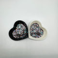 Two heart shaped containers, one black, one white, containing a Blue/Black/Light Teal/Rose Gold/Light Pink chunky glitter mix.
