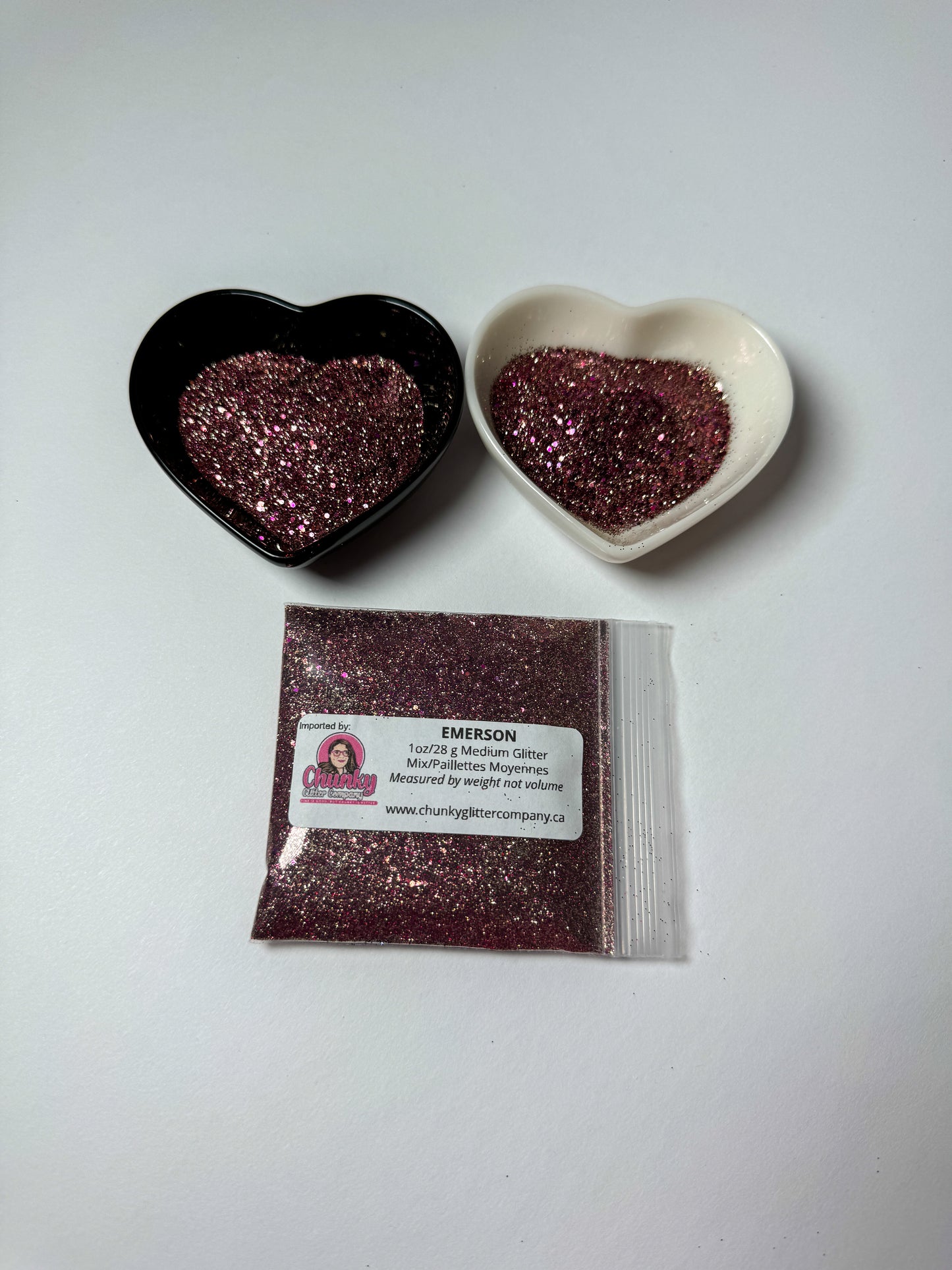 Two heart-shaped containers of glitter, one black and the other white, both showing an extra fine, maroon glitter mix and a bag of glitter with a label that reads Emerson.