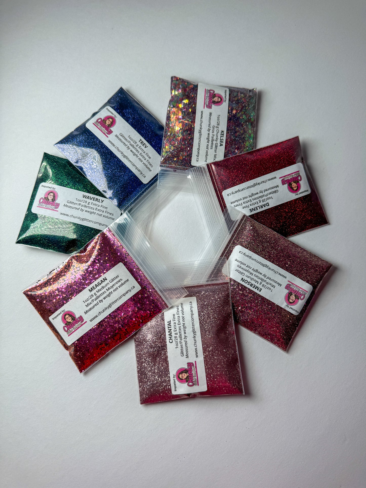 Seven packets of glitter in various colors arranged in a spiral pattern on a white background, each with a label indicating the glitter name.