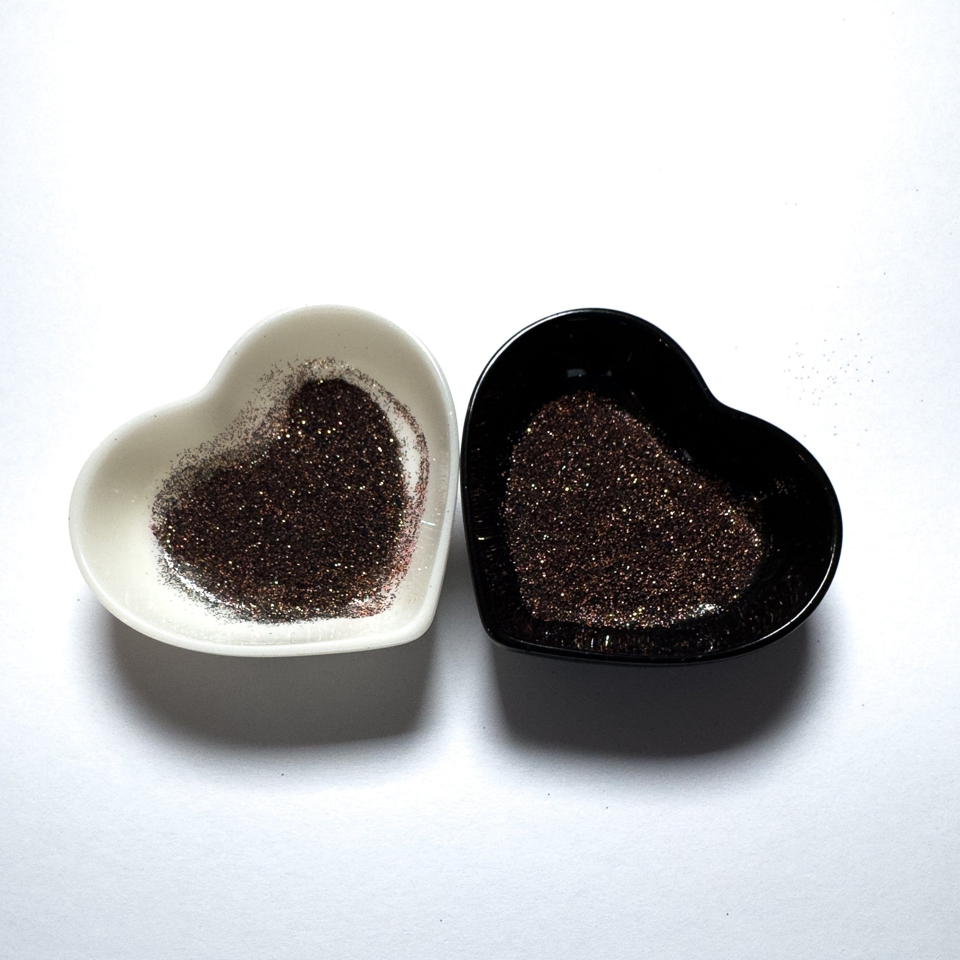 Two heart-shaped containers of glitter, one black and the other white, both showing an extra fine, multicolored glitter mix.