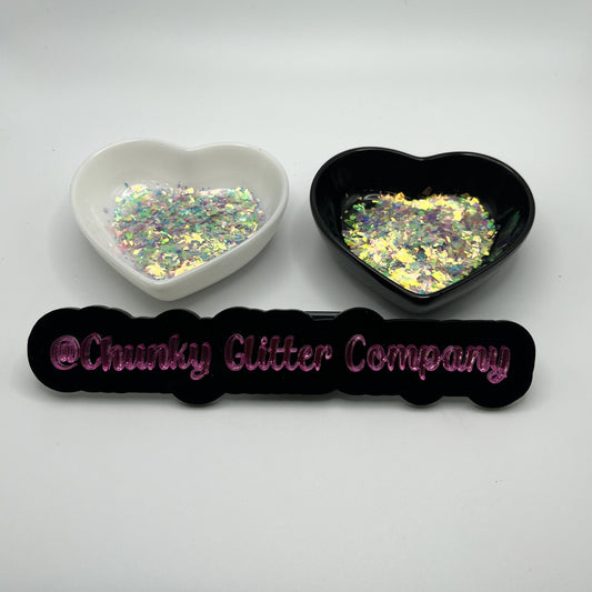 Two heart shaped containers, one black, one white, containing a Pink/Purple/Green/Orange chunky glitter mix, with a label in front that reads '@ChunkyGlitterCompany'.
