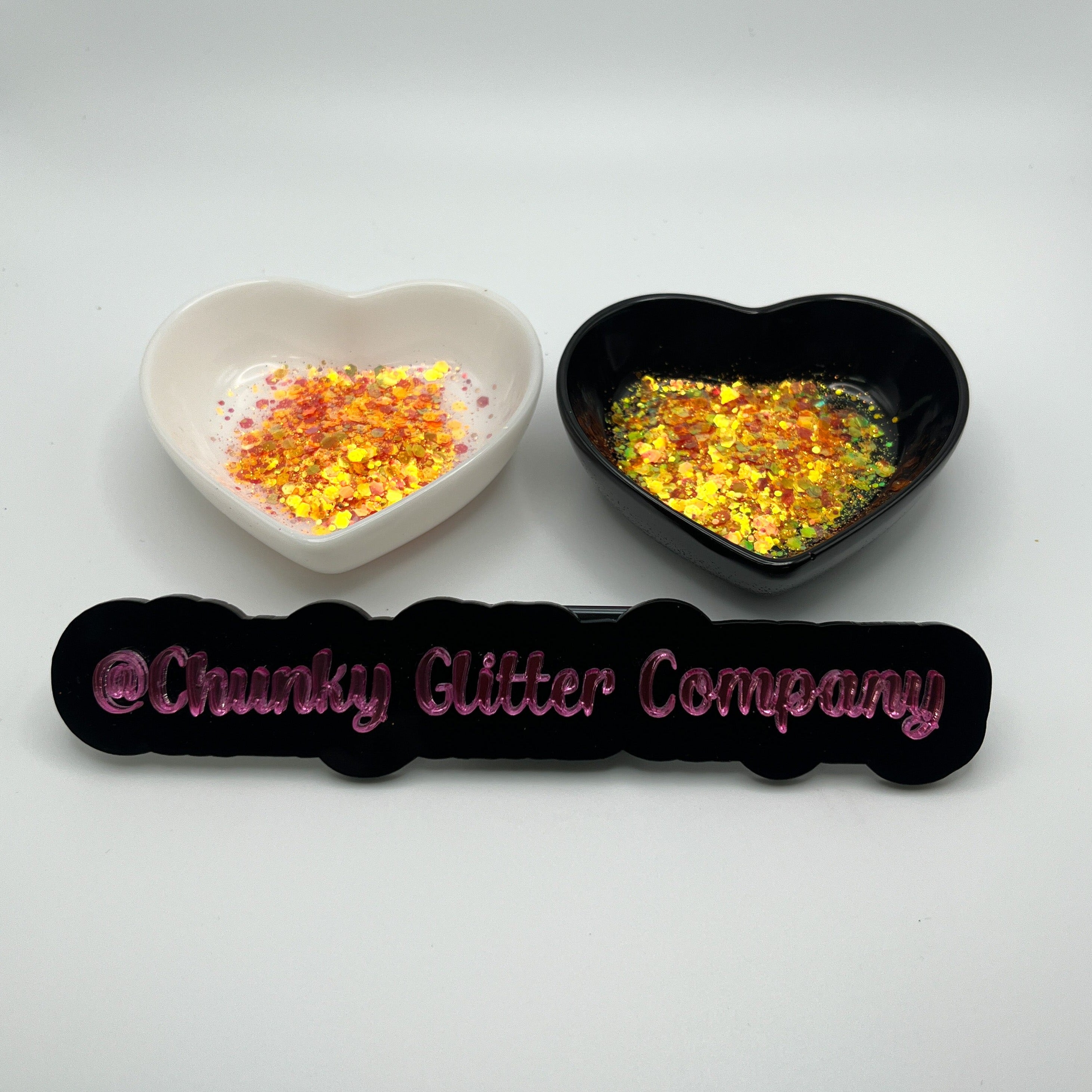 Two heart shaped containers, one black, one white, containing a Orange/Burnt Orange chunky glitter mix, with a label in front that reads '@ChunkyGlitterCompany'.