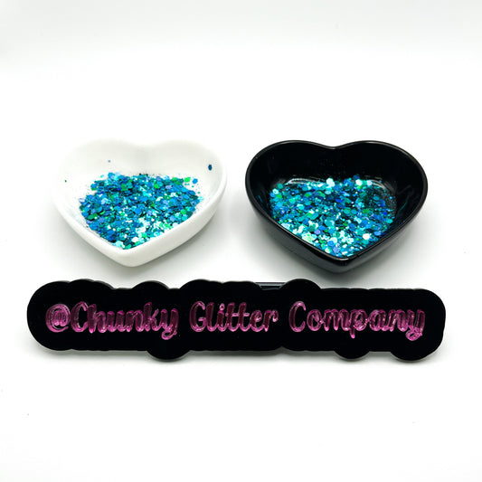 Two heart shaped containers, one black, one white, containing a Teal/Blue/Green/Hunter Green chunky glitter mix, with a label in front that reads '@ChunkyGlitterCompany'.