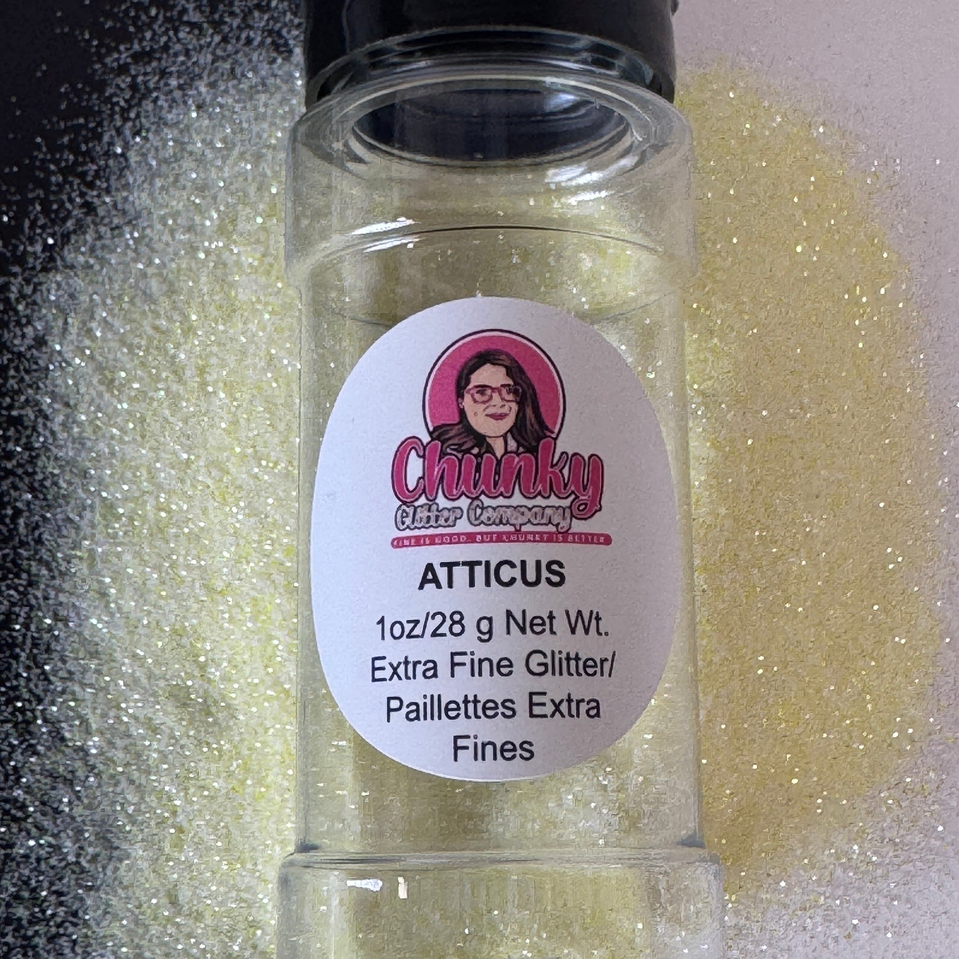 A clear plastic jar containing extra fine pastel yellow glitter with a label on the front showcasing the brand and product name, 'Atticus'.