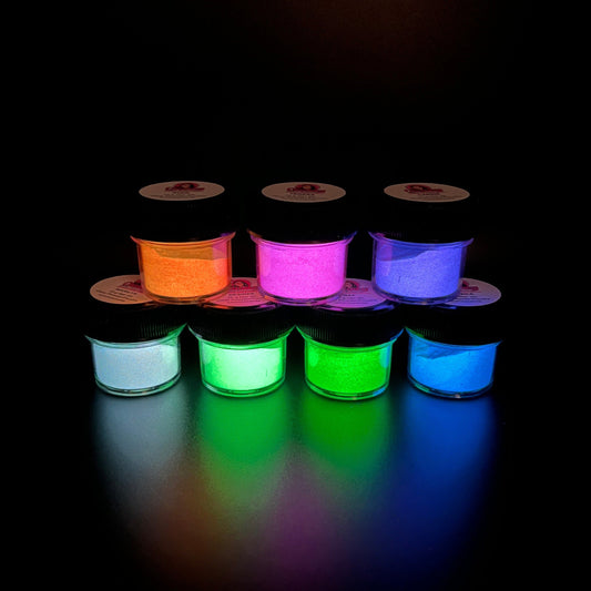 7 small jars of various colours of glow in the dark powders glowing on a black background