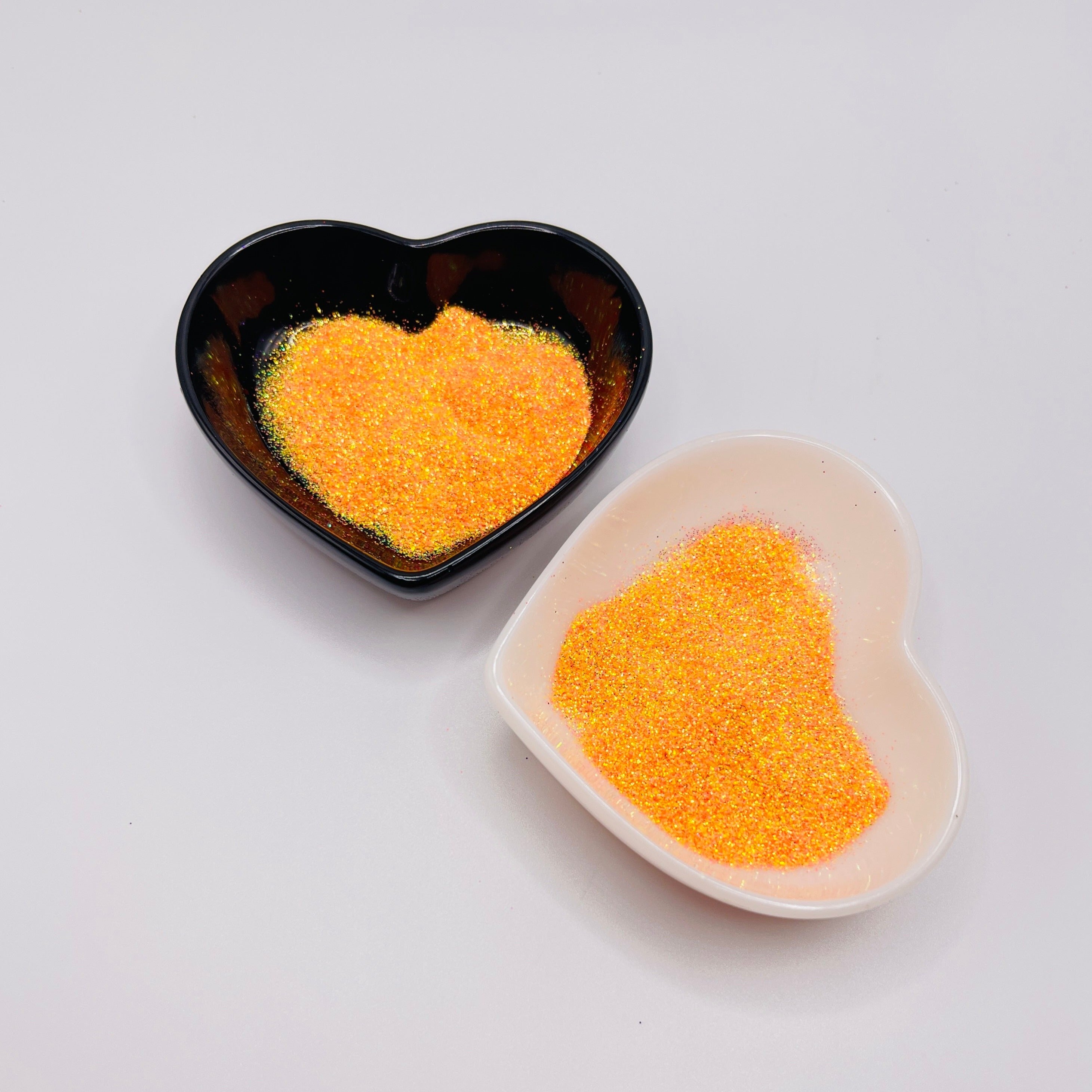 Two heart-shaped dishes containing orange-colored glitter, one black and one white.