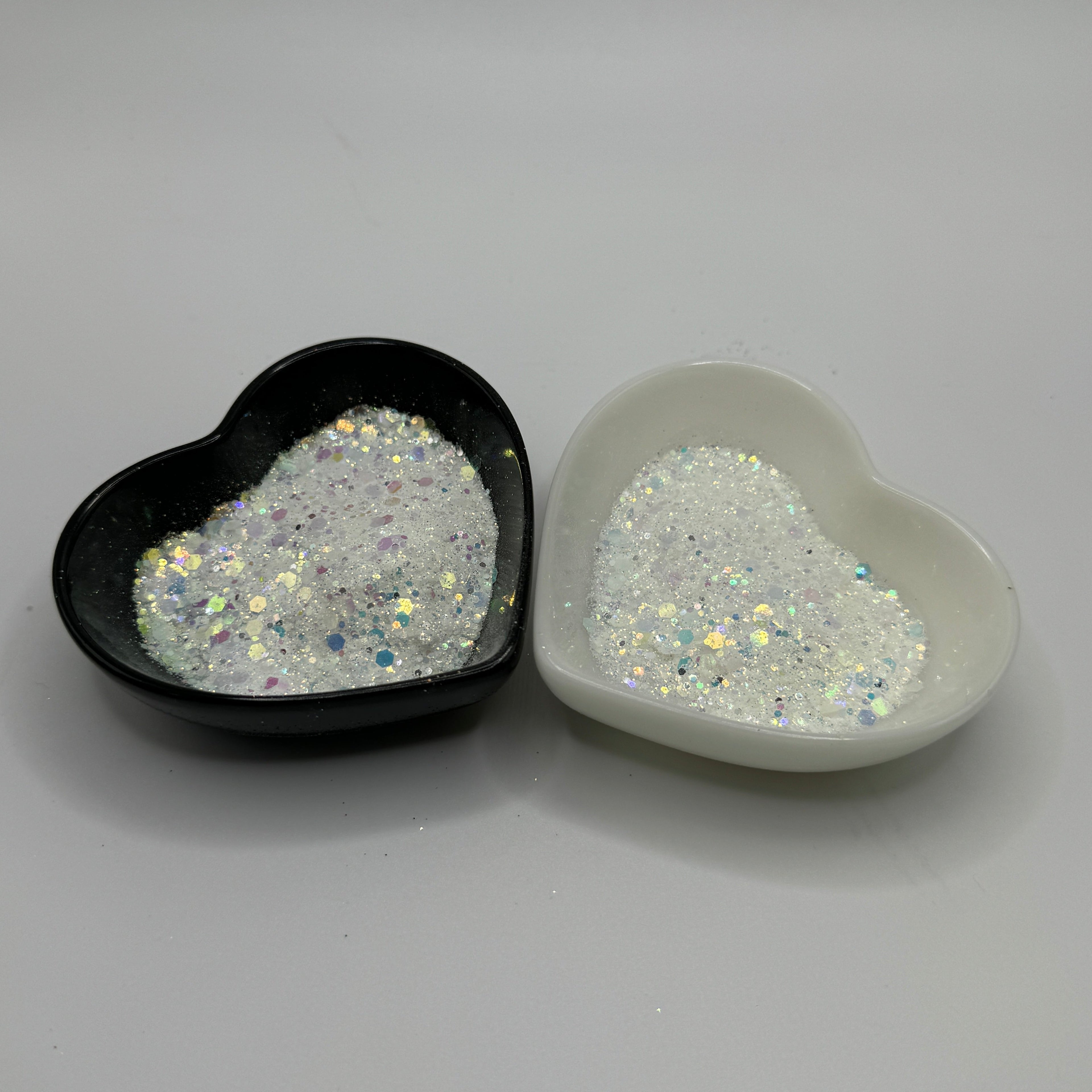 Two heart-shaped containers of glitter, one black and the other white, both showing a chunky white glitter mix