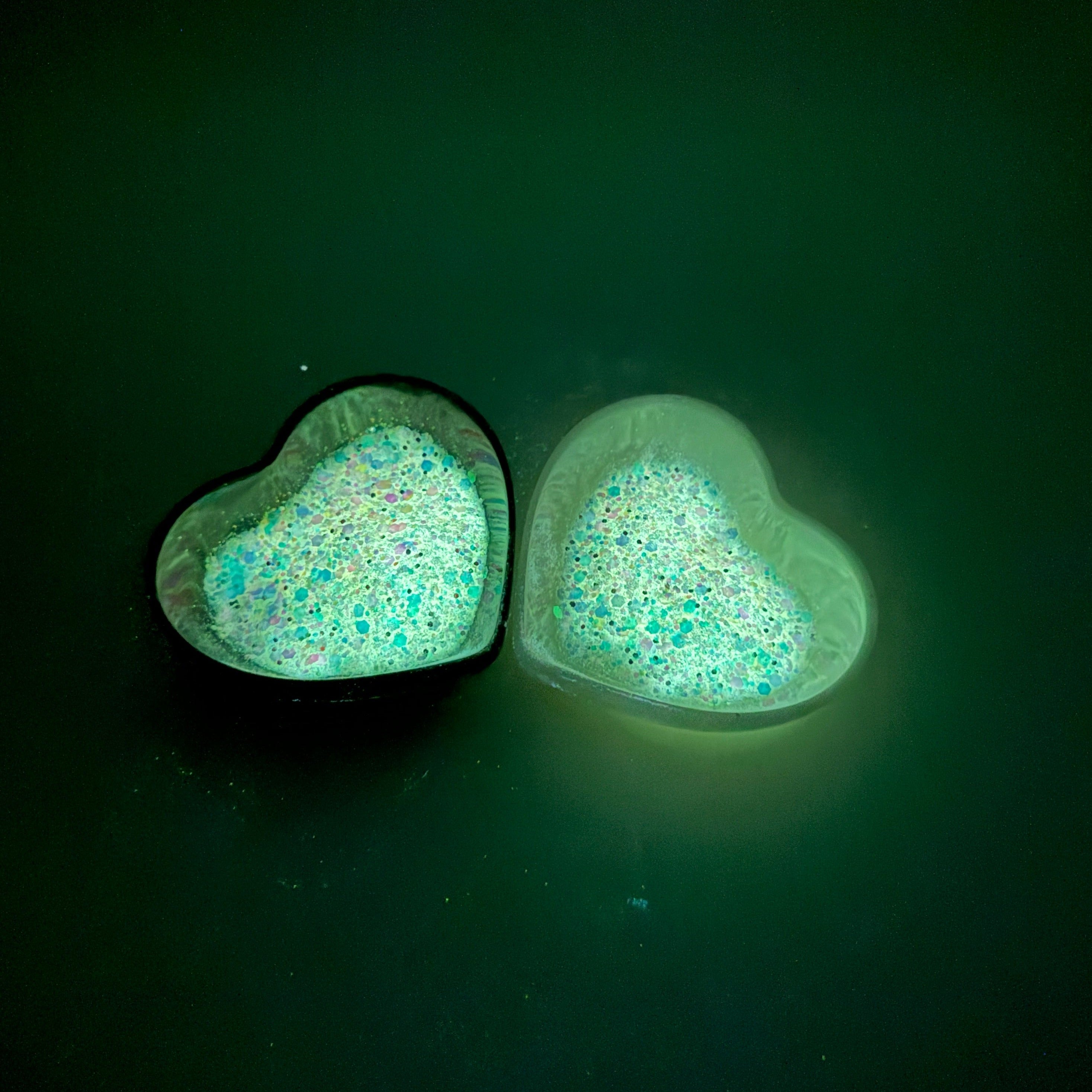 A photograph taken in the dark of two heart shaped containers, one black, one white, containing a White/Rainbow glow in the dark chunky glitter mix.