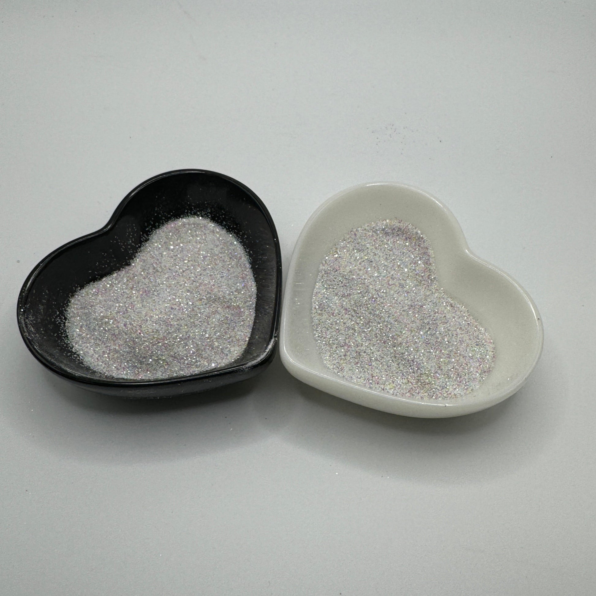 Two heart-shaped containers of glitter, one black and the other white, both showing an extra fine, white rainbow glitter mix