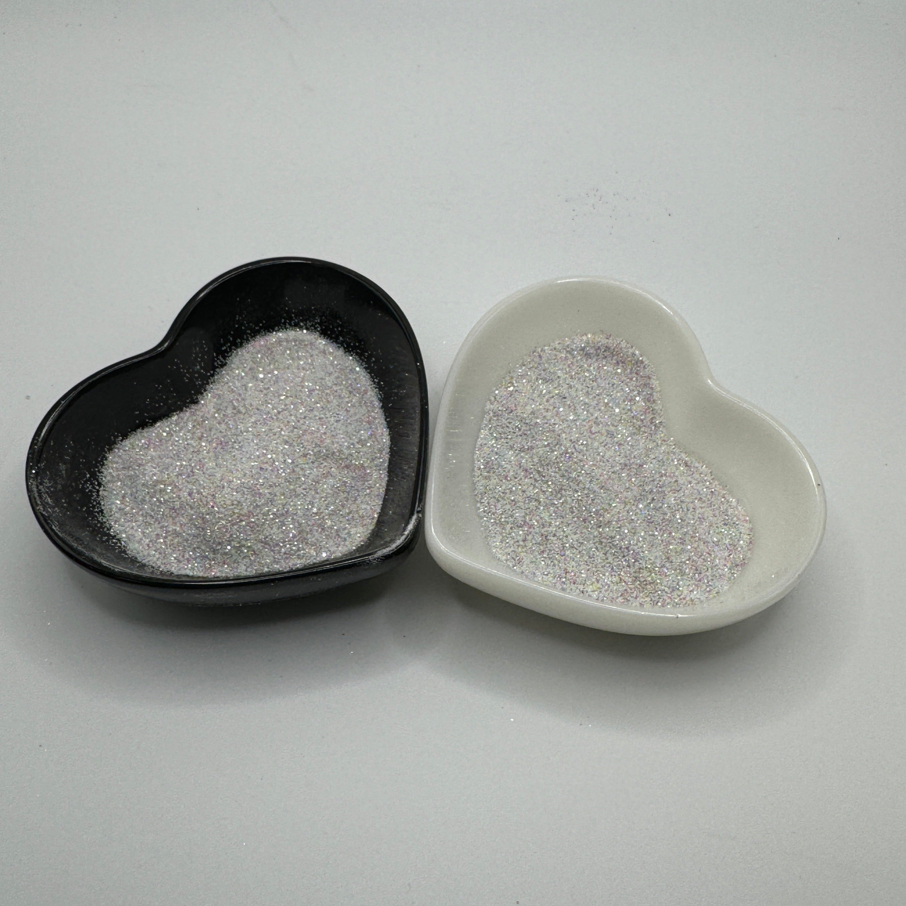 Two heart-shaped containers of glitter, one black and the other white, both showing an extra fine, white rainbow glitter mix