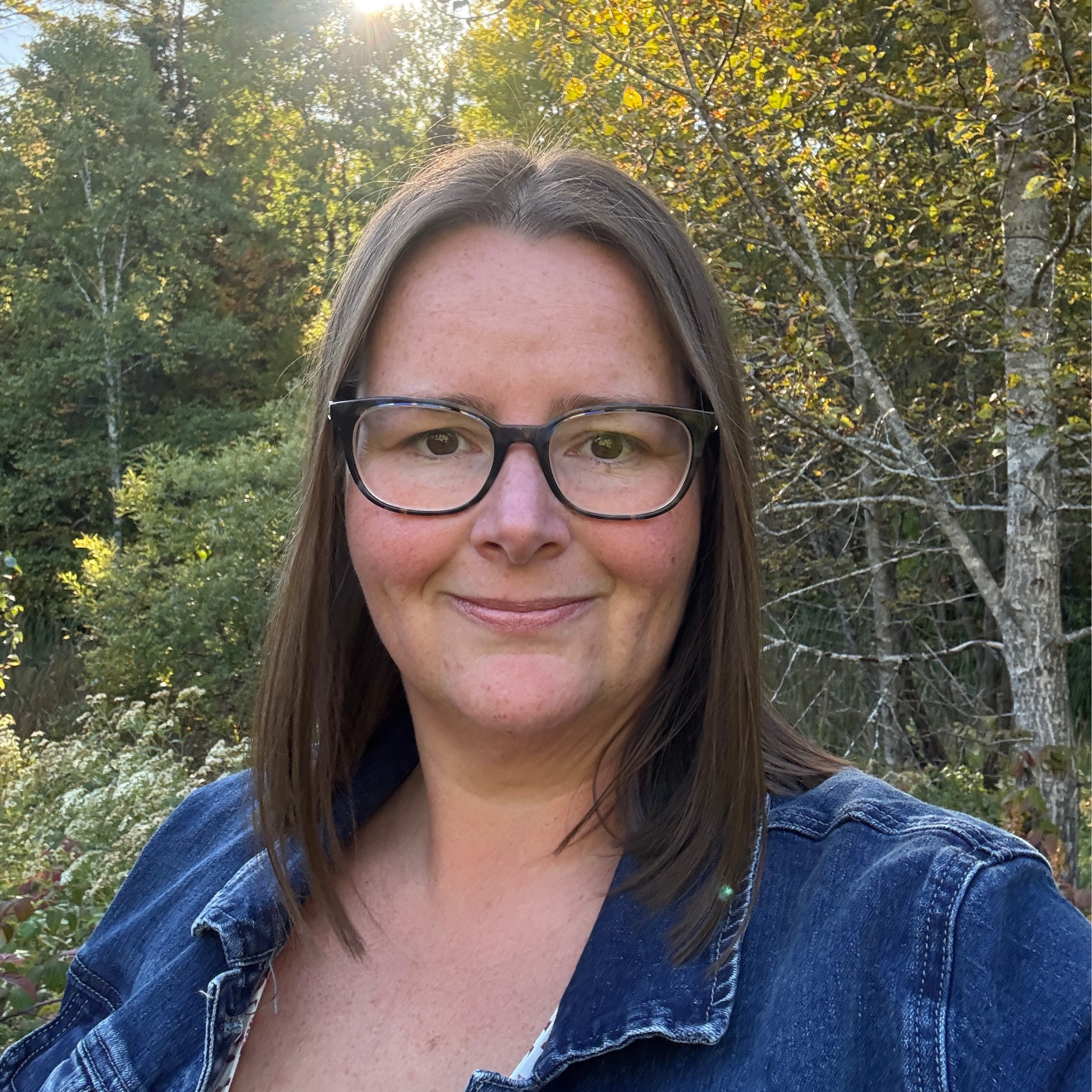 Kristen Butryn Owner wearing glasses and a denim jacket standing in a forest