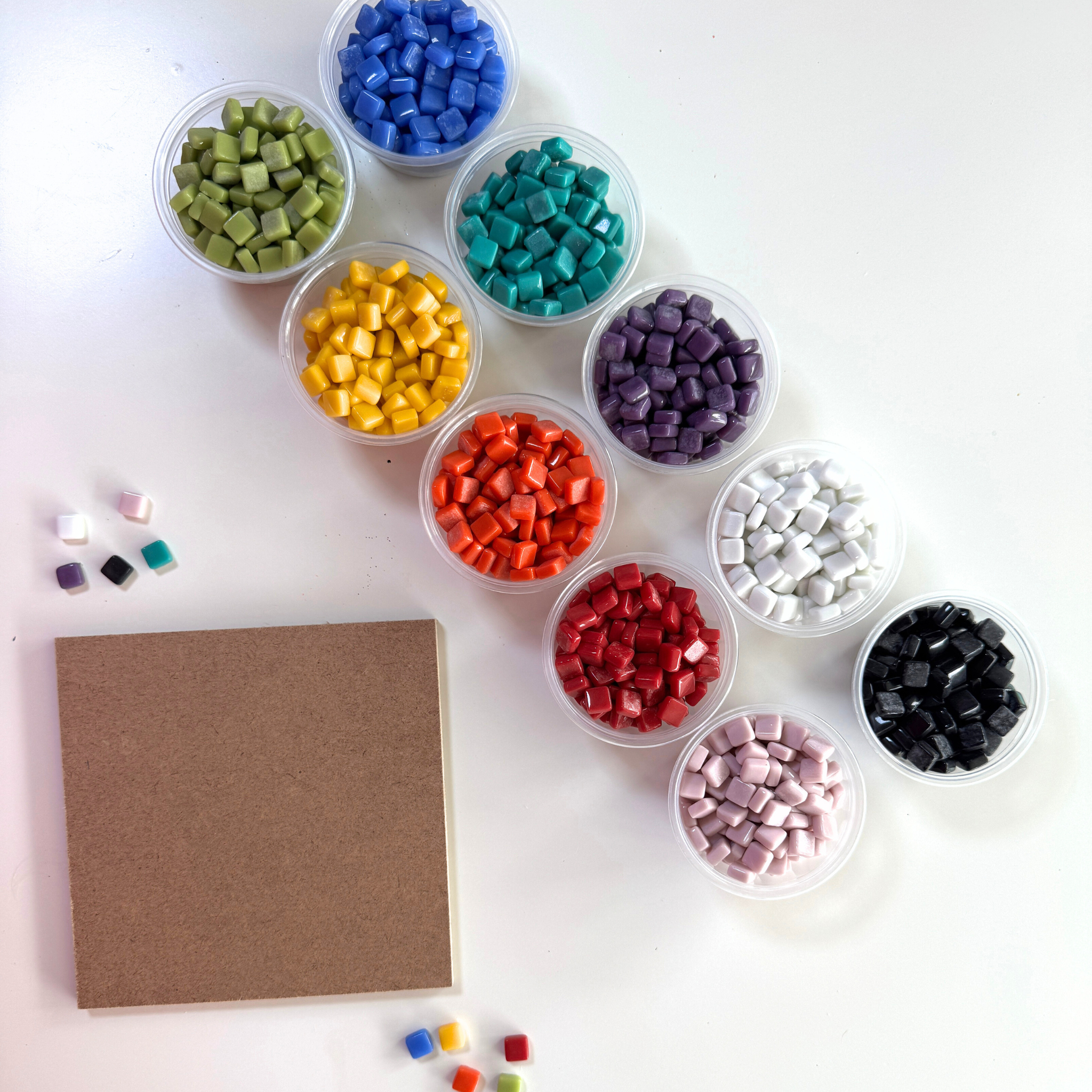 Assorted coloured tiny tiles in small containers on a white surface with a wooden coaster square.