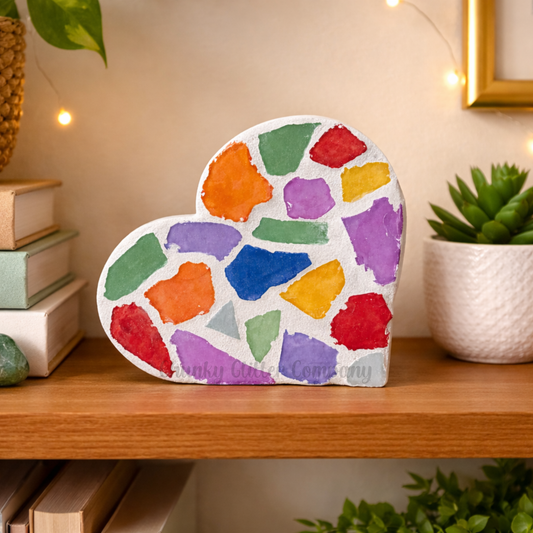 Colorful heart-shaped decorative item on a wooden shelf with plants and books in the background.