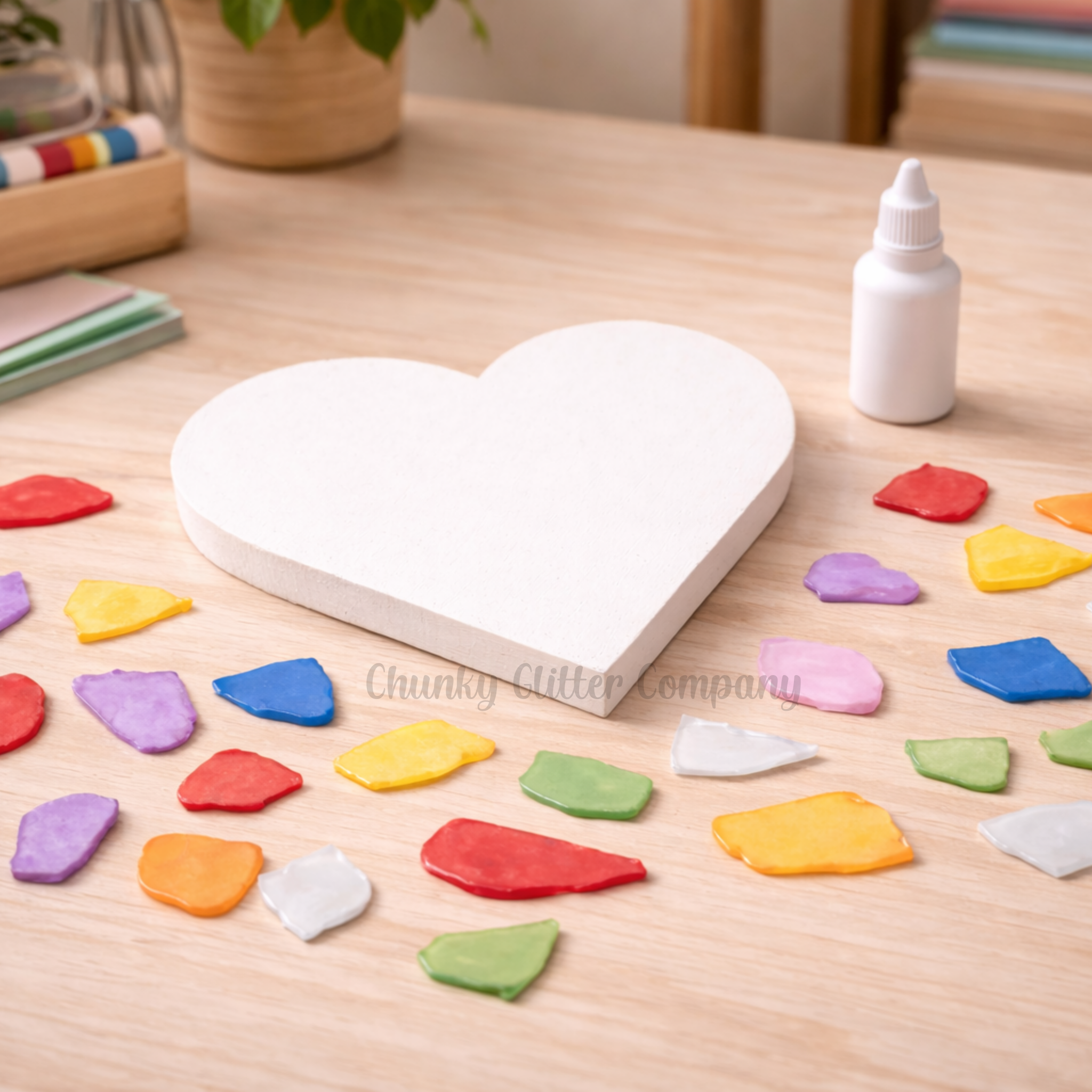 Heart-shaped white board with colorful beach glass on a wooden table, surrounded by a bottle of glue and a plant.